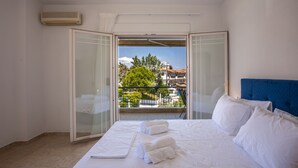 Una camera d'albergo con un letto, lenzuola bianche, un portatile e un balcone con vista su edifici e alberi.