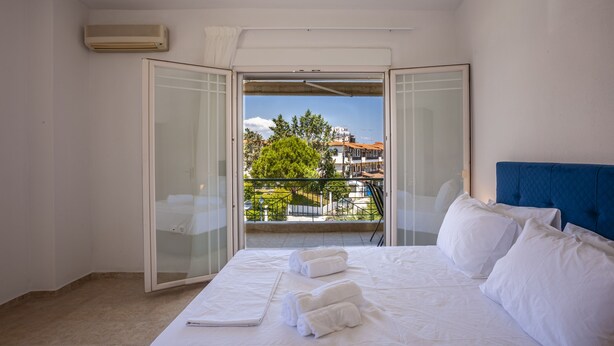 A hotel room with a bed, white linens, a laptop, and a balcony with a view of buildings and trees. - Aris Luxury Apartments (Kassandra)