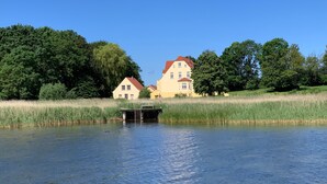 A calm lake with a dock, surrounded by lush greenery and two houses with red roofs. - Holiday Apartment "Gut Grubnow Rügen - 3" with Lake View, Shared Terrace & Wi-Fi (Neuenkirchen)