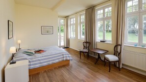 A bedroom with a bed, bedside lamps, a chair, a small table, and large windows with curtains. - Holiday Apartment "Gut Grubnow Rügen - 3" with Lake View, Shared Terrace & Wi-Fi (Neuenkirchen)