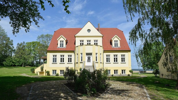 A large, yellow building with a red roof, a central entrance, and a cobblestone driveway. - Holiday Apartment "Gut Grubnow Rügen - 3" with Lake View, Shared Terrace & Wi-Fi (Neuenkirchen)