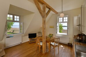 A room with a wooden dining table, chairs, a television, and a view of the sea through large windows.