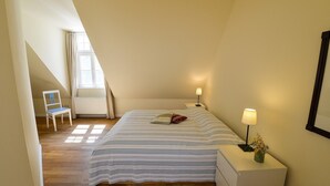 Un dormitorio con techo inclinado, una cama con ropa de cama a rayas, una silla, lámparas de noche y una mesita con un florero.