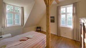 A bedroom with a sloped ceiling, a bed with a striped bedspread, two windows with curtains, a radiator, and a small table. - Apartment 'Gut Grubnow Rügen - Ferienwohnung 7' with Lake View, Shared Garden and Wi-Fi (Neuenkirchen)