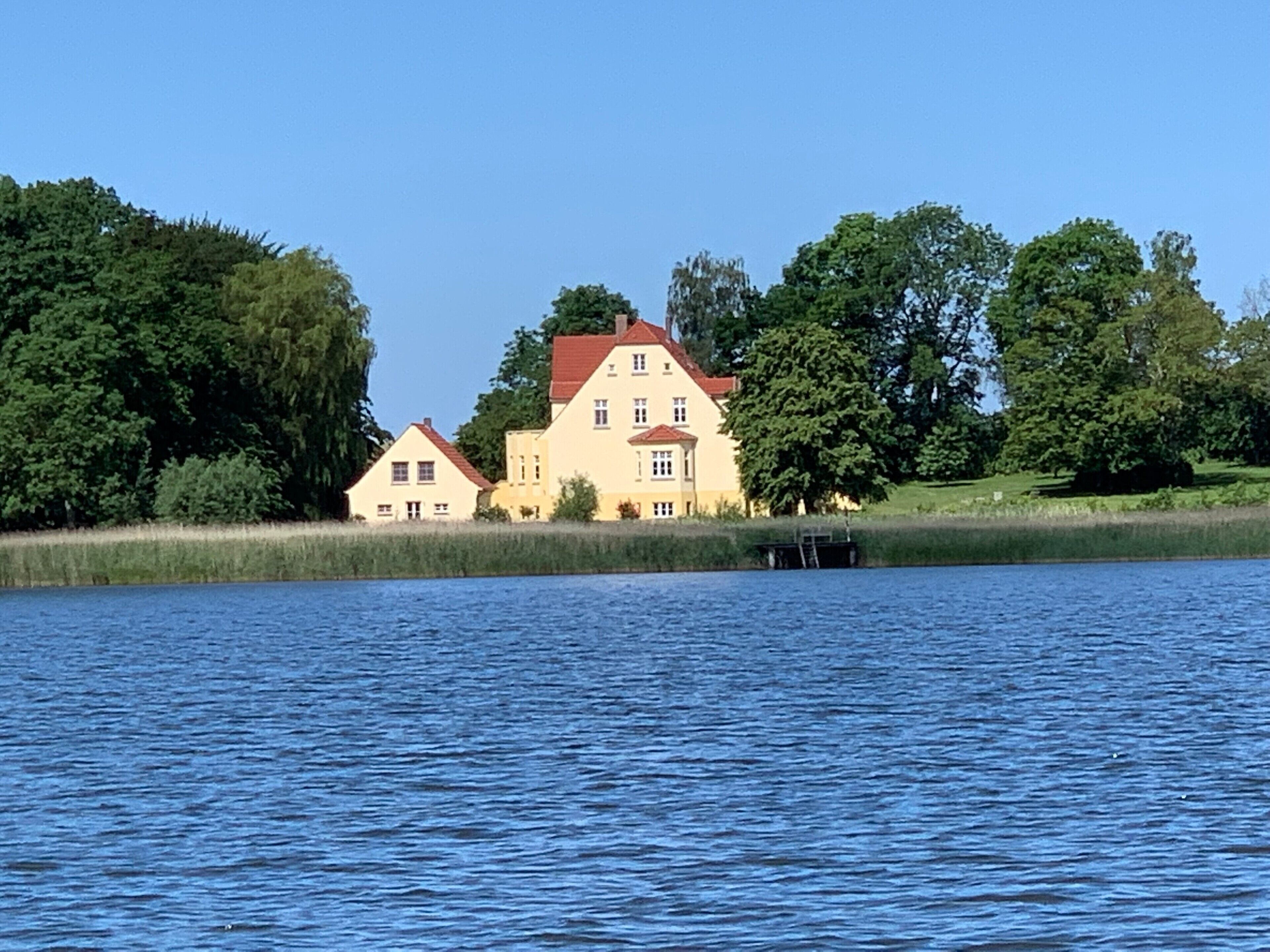 A large house with a red roof by a lake, surrounded by trees.