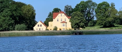 A large house with a red roof by a lake, surrounded by trees.