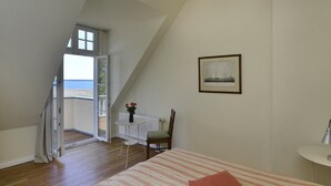 Une chambre à coucher avec un lit, une chaise, une table ornée de fleurs, un tableau encadré et une vue sur la mer par la fenêtre.