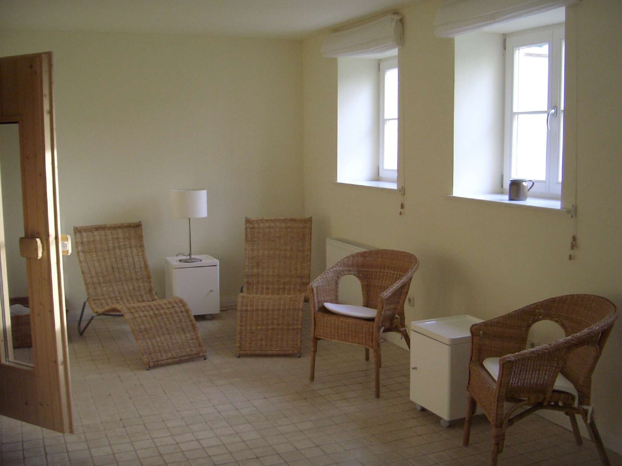 A room with wicker chairs, a small white cabinet, and a lamp. There are windows allowing natural light.