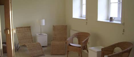 A room with wicker chairs, a small white cabinet, and a lamp. There are windows allowing natural light.