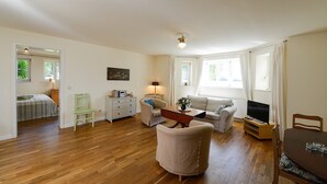 A spacious living room with a sofa, a dining table, and a TV. There is a window with curtains, a painting on the wall, and a wooden floor. - Holiday Apartment "Gut Grubnow Rügen - 1" with Lake View, Shared Garden & Wi-Fi (Neuenkirchen)