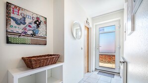 En gang med en fletkurv på et hvidt konsolbord, et spejl og en dør der fører til en balkon.