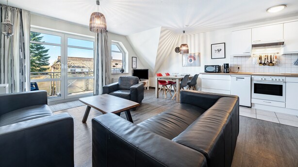 En moderne stue med sofa, sofabord og spiseområde med røde stole. Et køkken med hvide skabe og hårde hvidevarer er synligt.