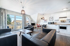 Un salon moderne avec un canapé, une table basse et un coin repas avec des chaises rouges. On peut apercevoir une cuisine avec des meubles blancs et des appareils électroménagers.