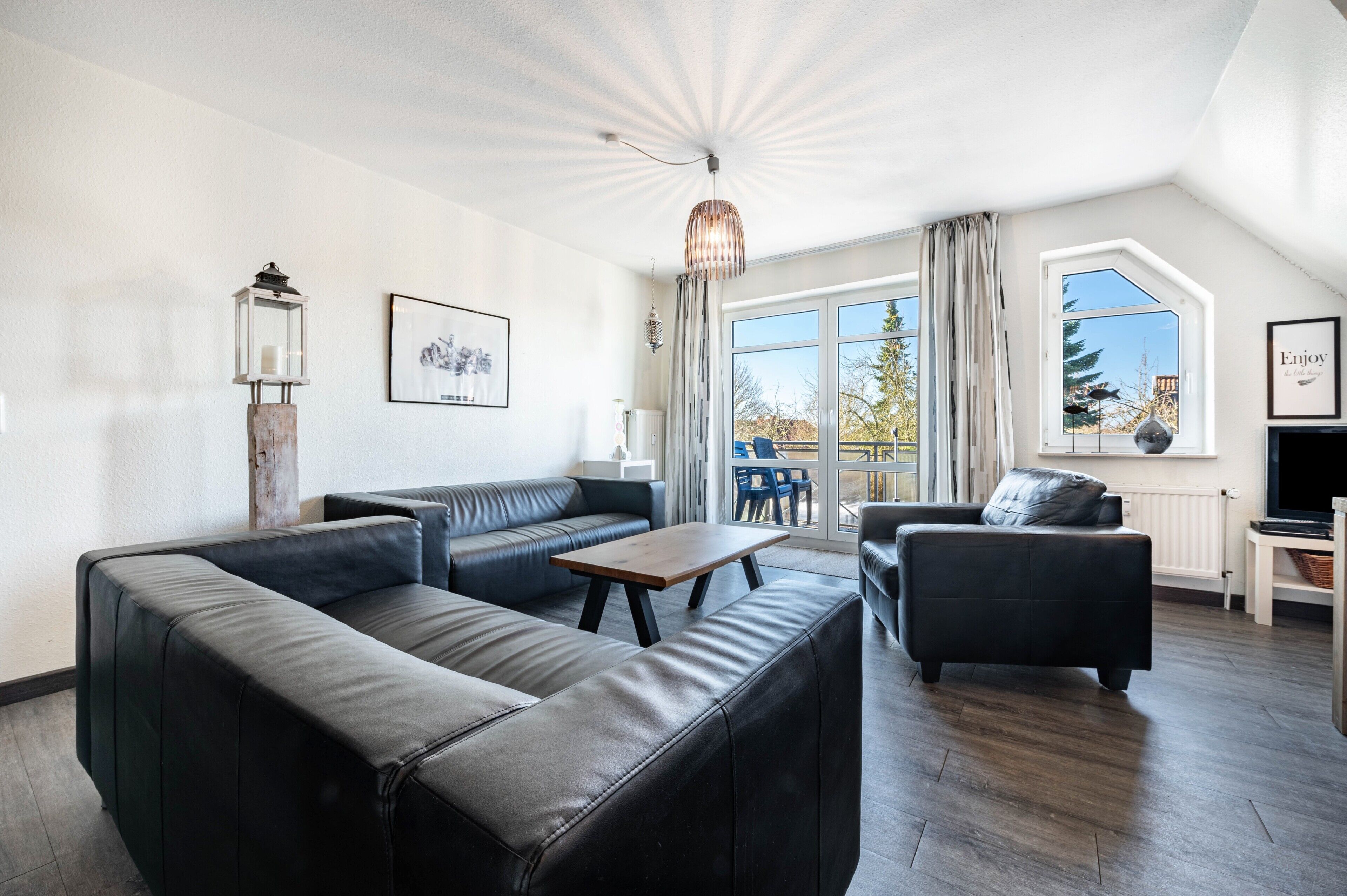 A modern living room with a sectional sofa, a coffee table, and a TV. There are large windows with curtains, a ceiling fan, and a hanging light fixture.