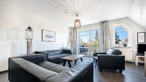 En moderne stue med en hjørnesofa, et sofabord og et fjernsyn. Store vinduer med gardiner, en loftsvifte og en hængende lampe.