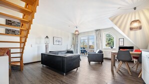 En moderne stue med en sort sofa, et spisebord med stole, en trappe og et stort vindue.