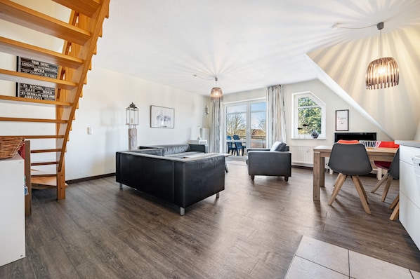 Un salon moderne avec un canapé noir, une table à manger avec des chaises, un escalier et une grande fenêtre.