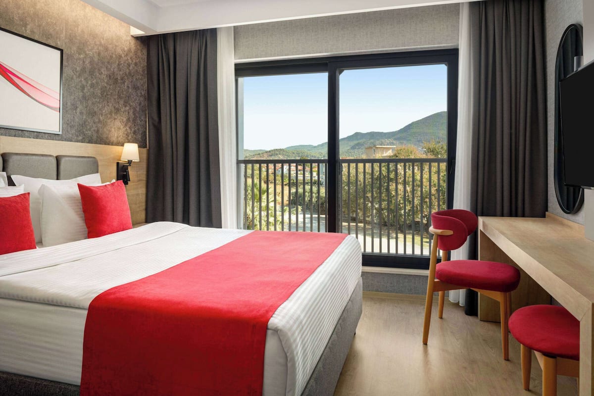 Een hotelkamer met een groot bed, een bureau en een stoel. Er is een balkon met uitzicht op de bergen.