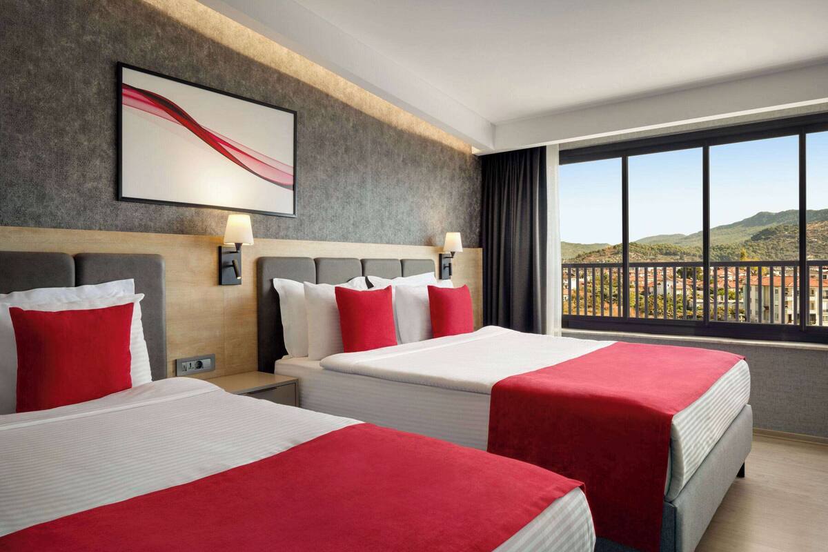 Een hotelkamer met twee bedden, een groot raam met uitzicht op heuvels en een modern abstract schilderij aan de muur.