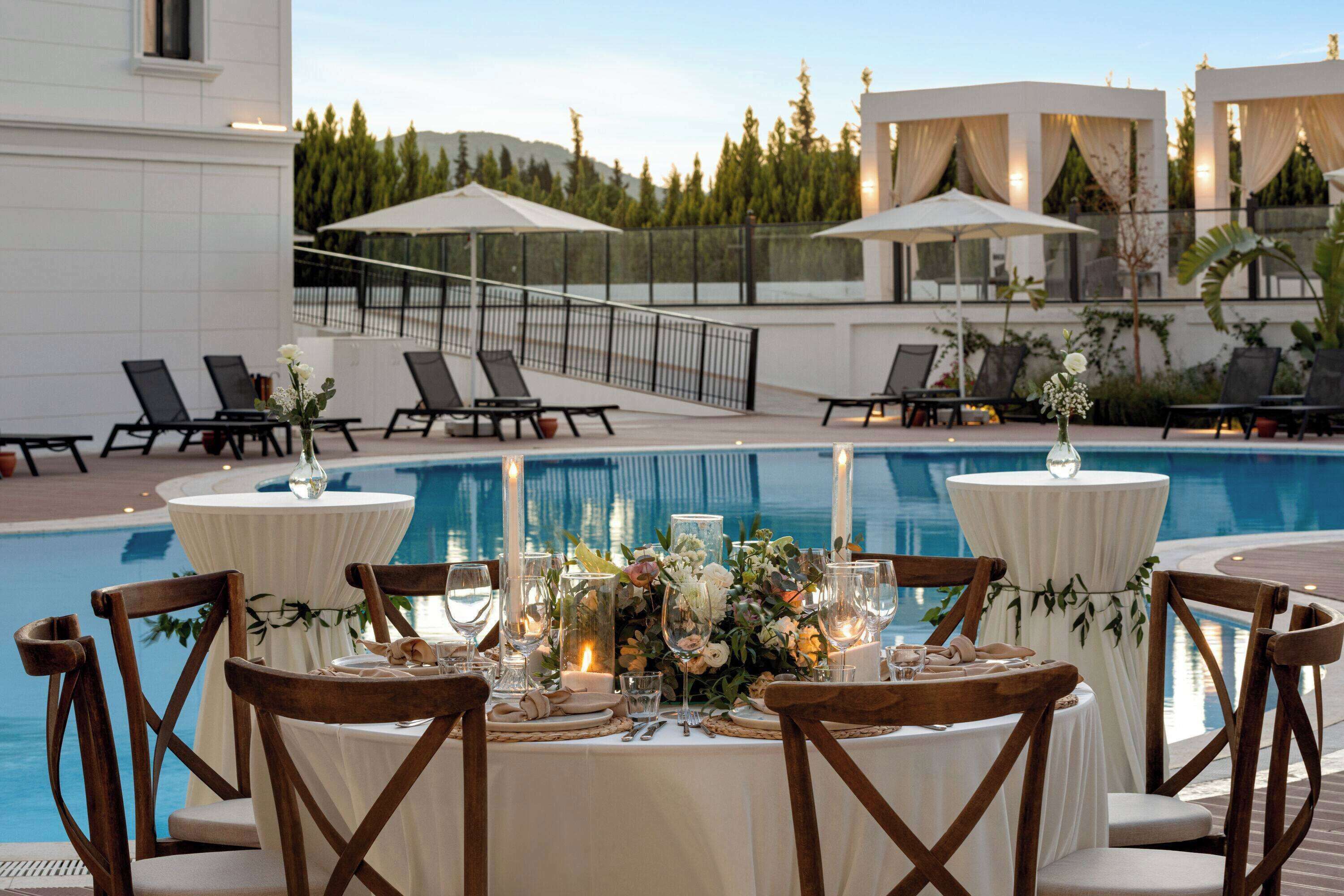 A poolside dining area with tables set for two, surrounded by lounge chairs, and overlooking a swimming pool.