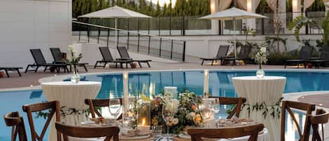 A poolside dining area with tables set for two, surrounded by lounge chairs, and overlooking a swimming pool.