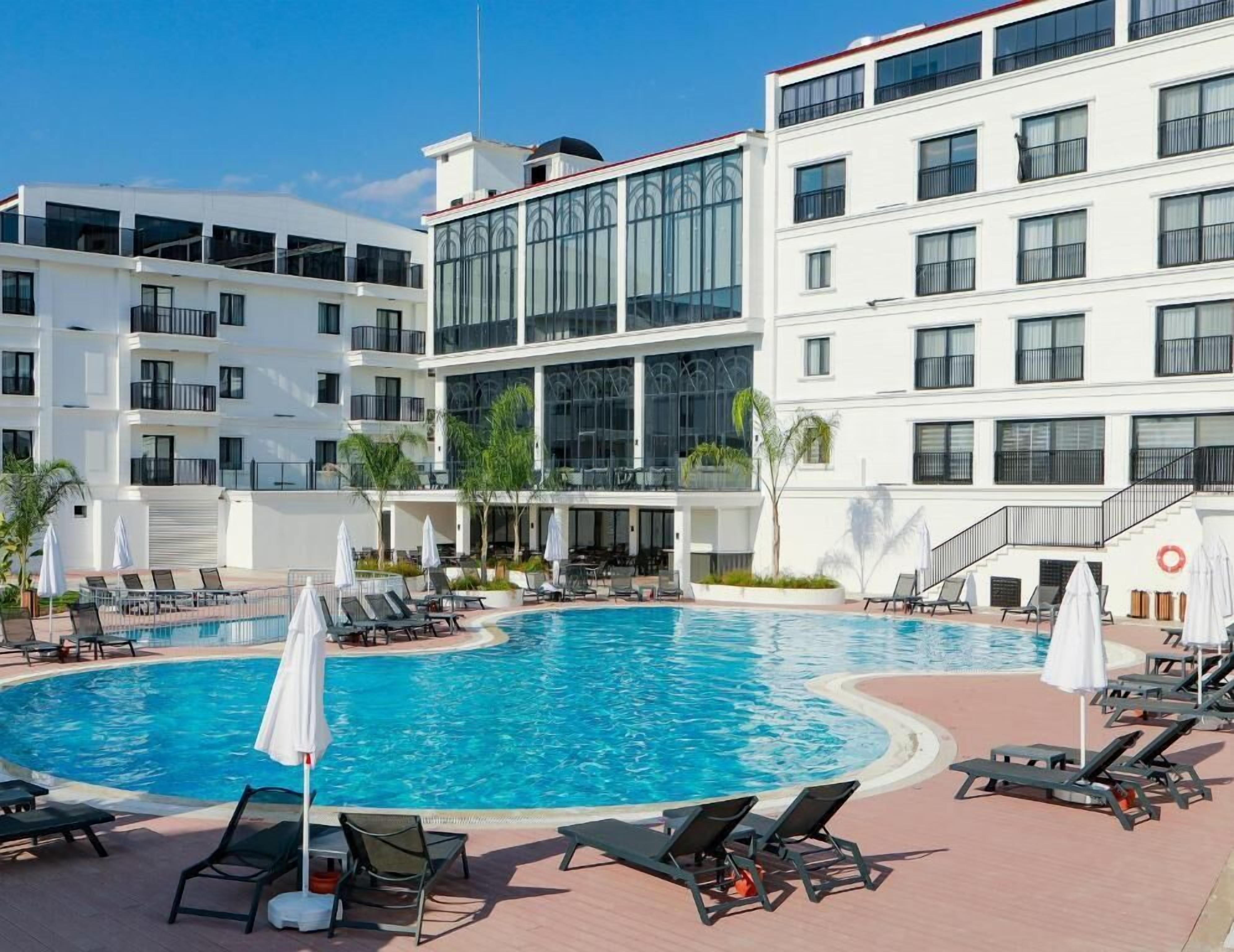 Ein modernes Hotel mit einem zentral gelegenen Schwimmbad, umgeben von Liegestühlen und Sonnenschirmen, und mehreren Balkonen mit Glasgeländern.