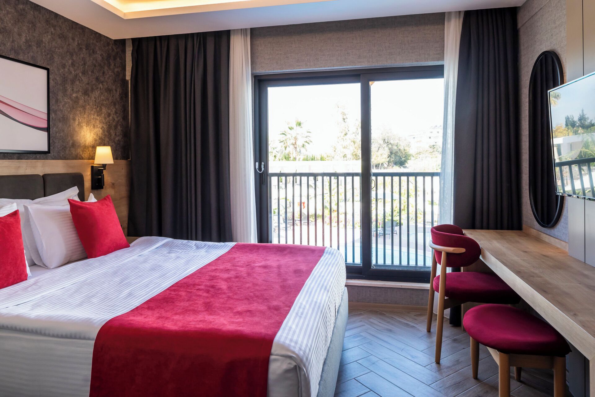 Ein Hotelzimmer mit einem Bett, einem Schreibtisch und einem Sessel. Ein Balkon bietet Blick auf Bäume und Gebäude.