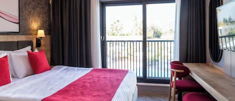 Habitación de hotel con cama, escritorio y silla. Cuenta con un balcón con vistas a árboles y edificios.