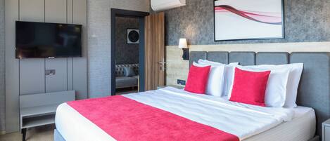 A modern hotel room with a large bed, a flat-screen TV, a wall-mounted air conditioner, and a grey sofa in the corner.