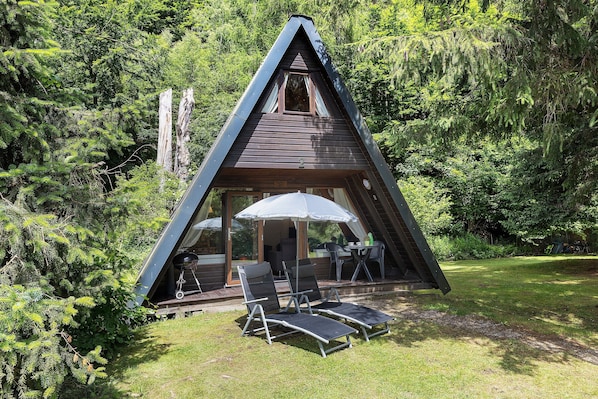 Une cabane triangulaire en bois dotée d’un balcon, de deux fauteuils et d’une table avec un parasol.