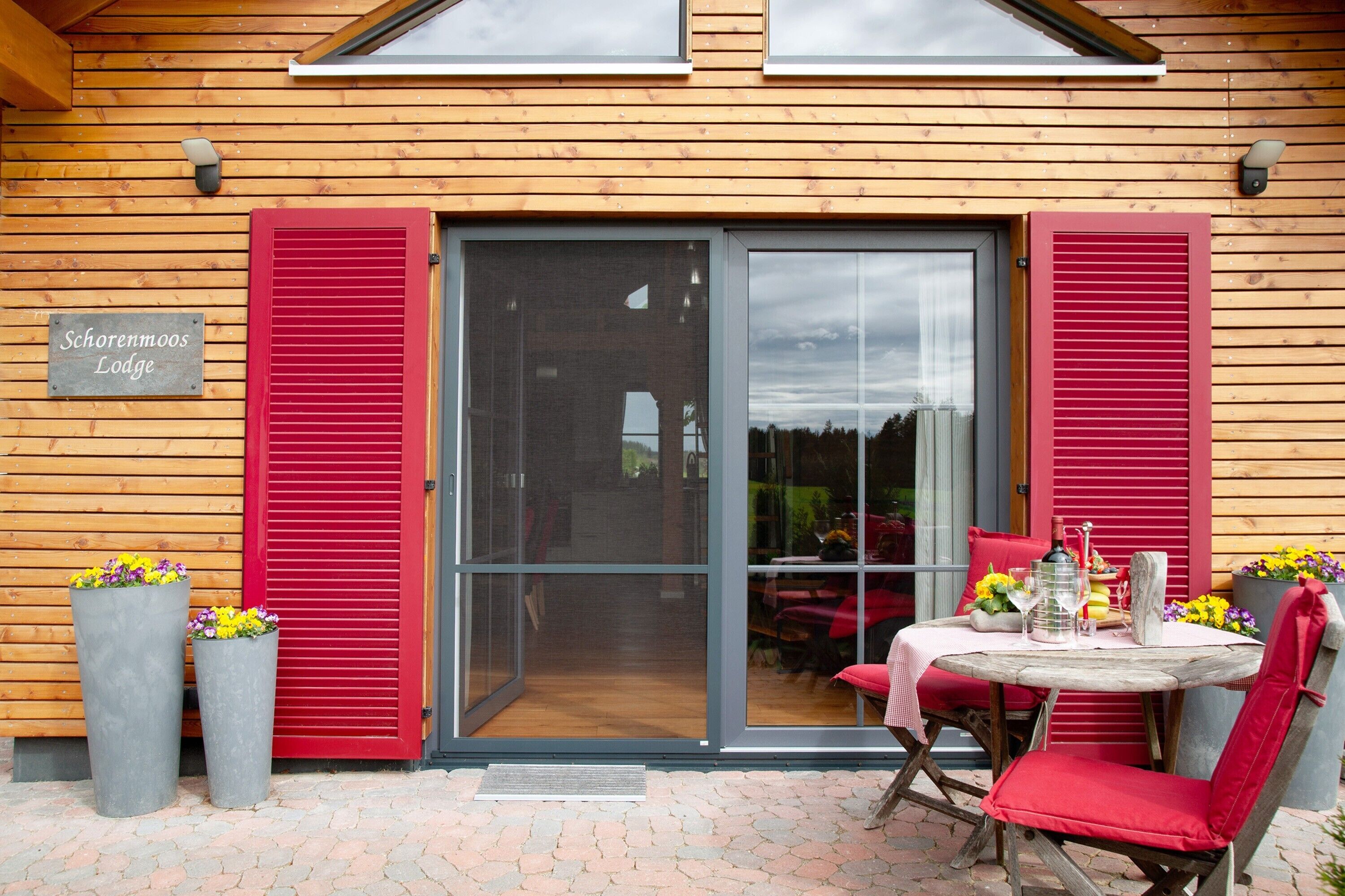 Een houten lodge met een schuifpui, rode luiken en een tafel met stoelen buiten.