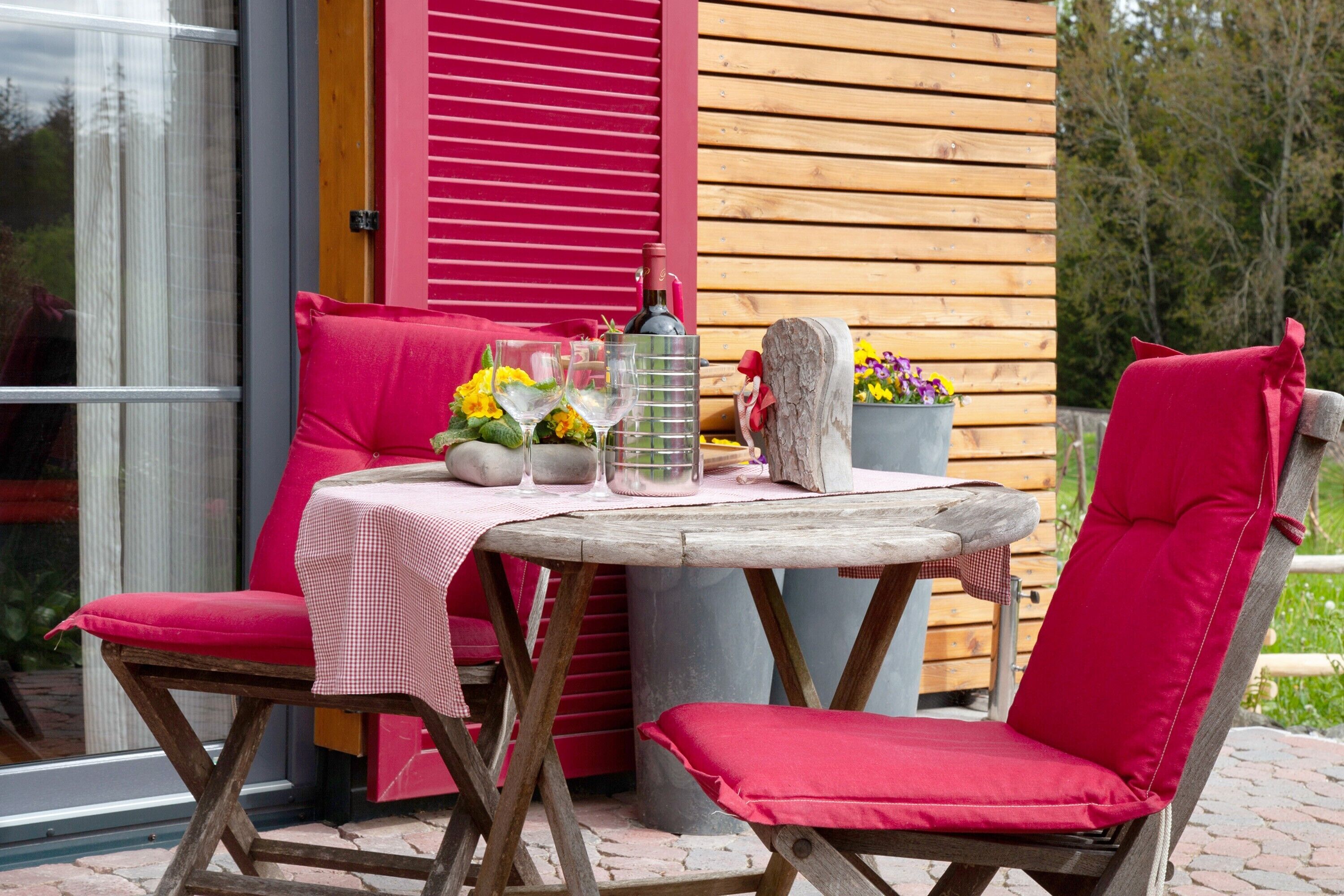 Una mesa de madera al aire libre con mantel a cuadros, dos sillas rojas acolchadas, una planta en maceta con flores y una botella de vino.