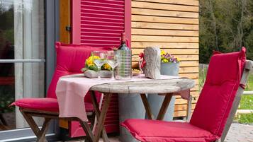 Una mesa de madera al aire libre con mantel a cuadros, dos sillas rojas acolchadas, una planta en maceta con flores y una botella de vino.