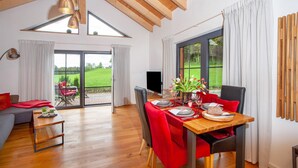 Een eetgedeelte met een tafel gedekt voor twee personen, een bank en een televisie in een kamer met houten balken en grote ramen.