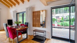 Een eetruimte met een houten tafel, rode stoelen, een bank en een groot raam met uitzicht op een tuin.