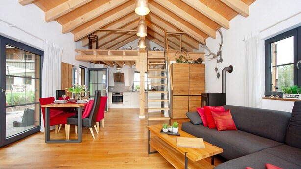 Een ruime woonkamer met een hoge plafond, houten balken, een eettafel met stoelen, een bank en uitzicht door grote ramen.