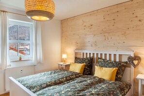 A bedroom with a wooden headboard, floral bedding, a bedside lamp, and a view of a building outside the window. - Holiday Apartment "Fuchsbau" with Mountain View, Balcony & Wi-Fi (Oberau)