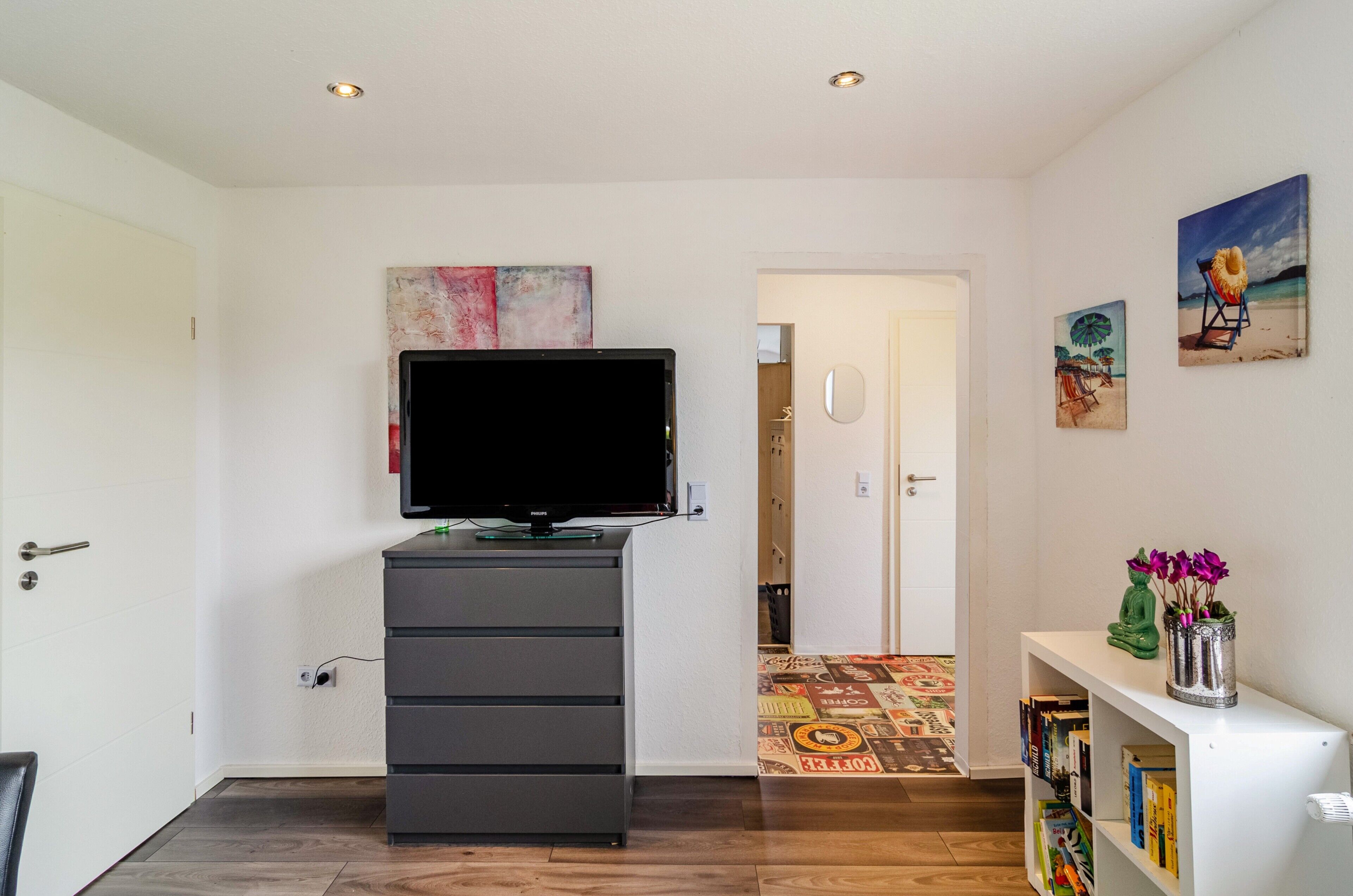 Ein Zimmer mit Fernseher auf einer Kommode, einem Bücherregal mit Büchern, einer Tür zu einem anderen Raum und einem Bild an der Wand.