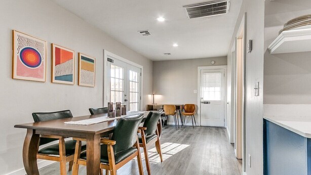 Un espace repas moderne avec une table et des chaises en bois, un coin cuisine à l’arrière-plan et des œuvres d’art accrochées au mur.