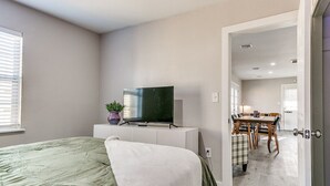 Une chambre à coucher avec un lit, un téléviseur posé sur un meuble, une table à manger avec des chaises, et une porte donnant sur une autre pièce.