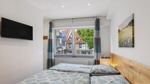Une chambre à coucher avec un lit, un téléviseur fixé au mur, une fenêtre avec des rideaux et un tableau accroché au mur.