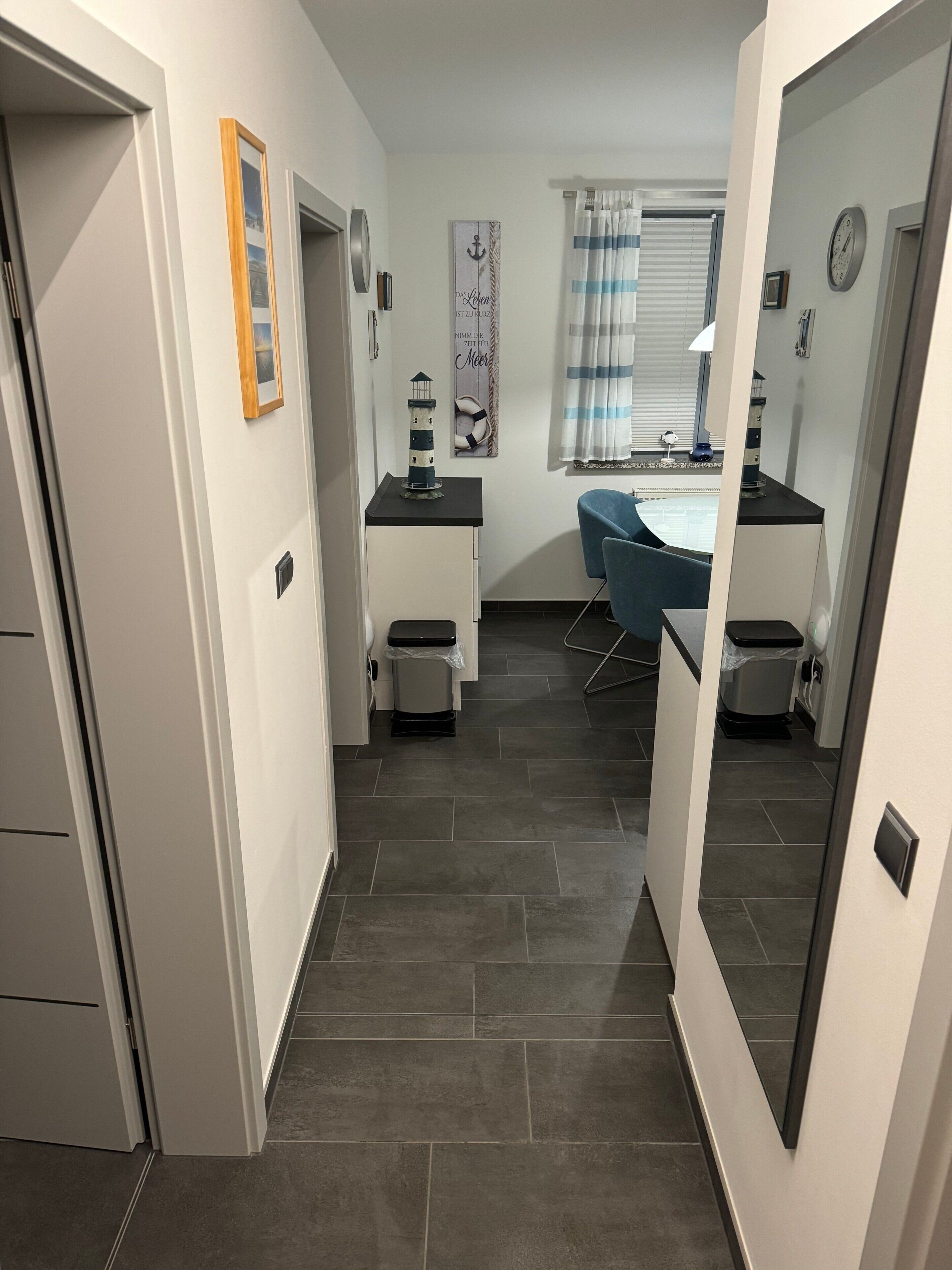 A narrow hallway with a small desk, a chair, a trash bin, and a window with curtains.
