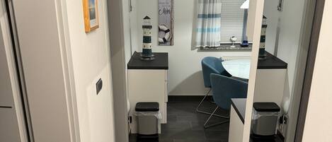 A narrow hallway with a small desk, a chair, a trash bin, and a window with curtains.