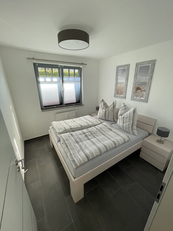 A bedroom with a bed, bedside tables, a window with blinds, a radiator, and framed pictures on the wall.