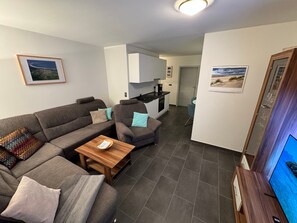 A modern living room with a sofa, armchair, and coffee table, a kitchen area, and a television.