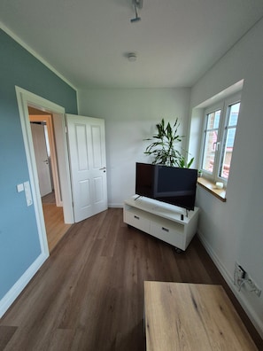 Ett rum med en platt-TV placerad på ett vitt TV-bord, en krukväxt, ett träbord och ett fönster med utsikt över en byggnad med tegelstensfasad.