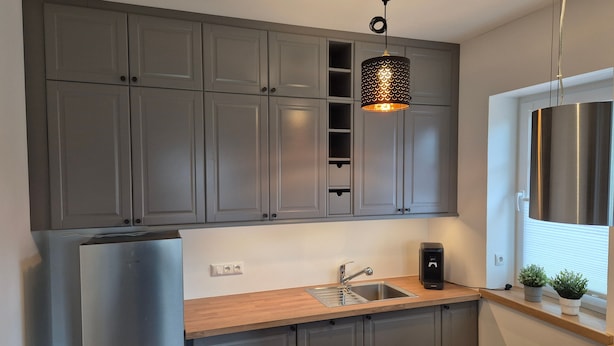 Une cuisine moderne avec des meubles gris, un réfrigérateur en acier inoxydable, un plan de travail en bois, un évier et un luminaire suspendu.