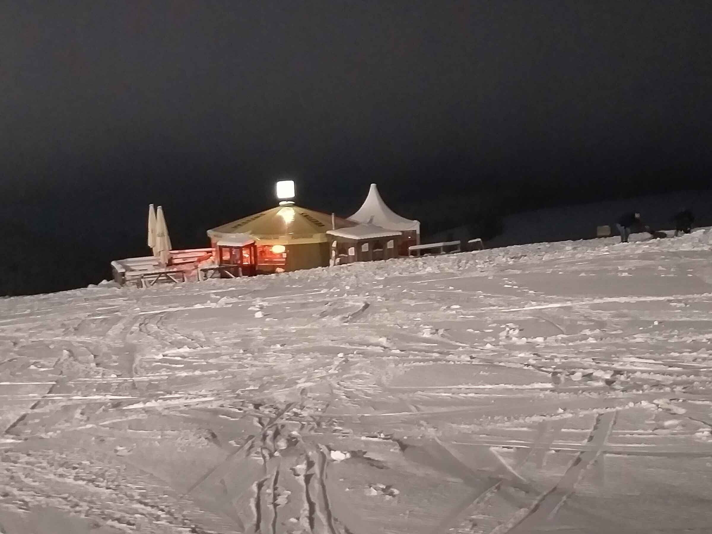 A snow-covered landscape with a small building featuring a lit sign, a tent, and a few chairs.