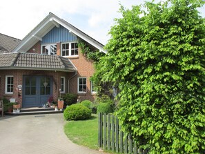 Une maison à deux étages avec une façade en briques, un porche à l’avant orné de plantes en pot et une clôture en bois.