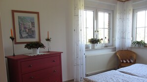 A bedroom with a red dresser, a bed with a floral bedspread, a wicker chair, and a window with curtains.